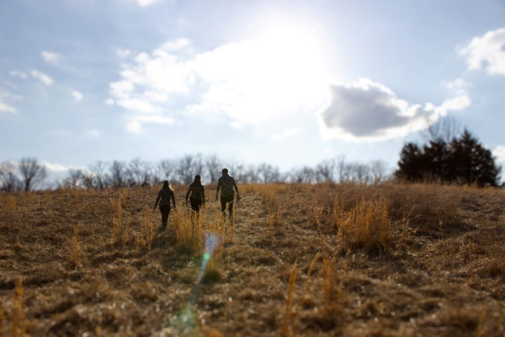 Three people on a guided hunting trip walk across an open field with the sun shining down.