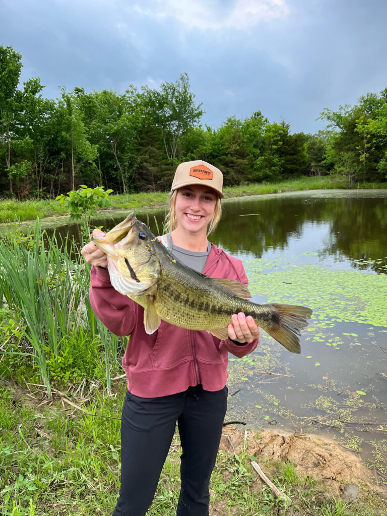 Young woman holds a fish up for a photo with a pond and trees behind her.