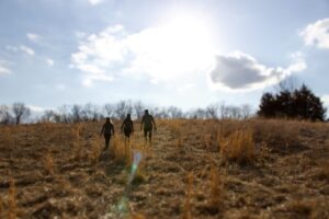 Three hunters walk across an open field with the sun shining overhead