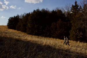 Two hunters walk up a hill in an open field surrounded by trees